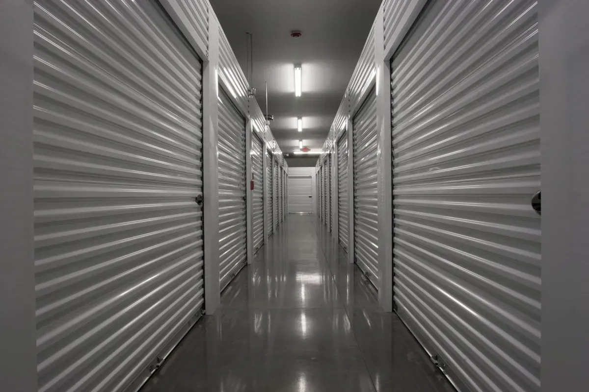 An indoor hallway of metal self storage units with closed roll-up doors and bright overhead lighting.